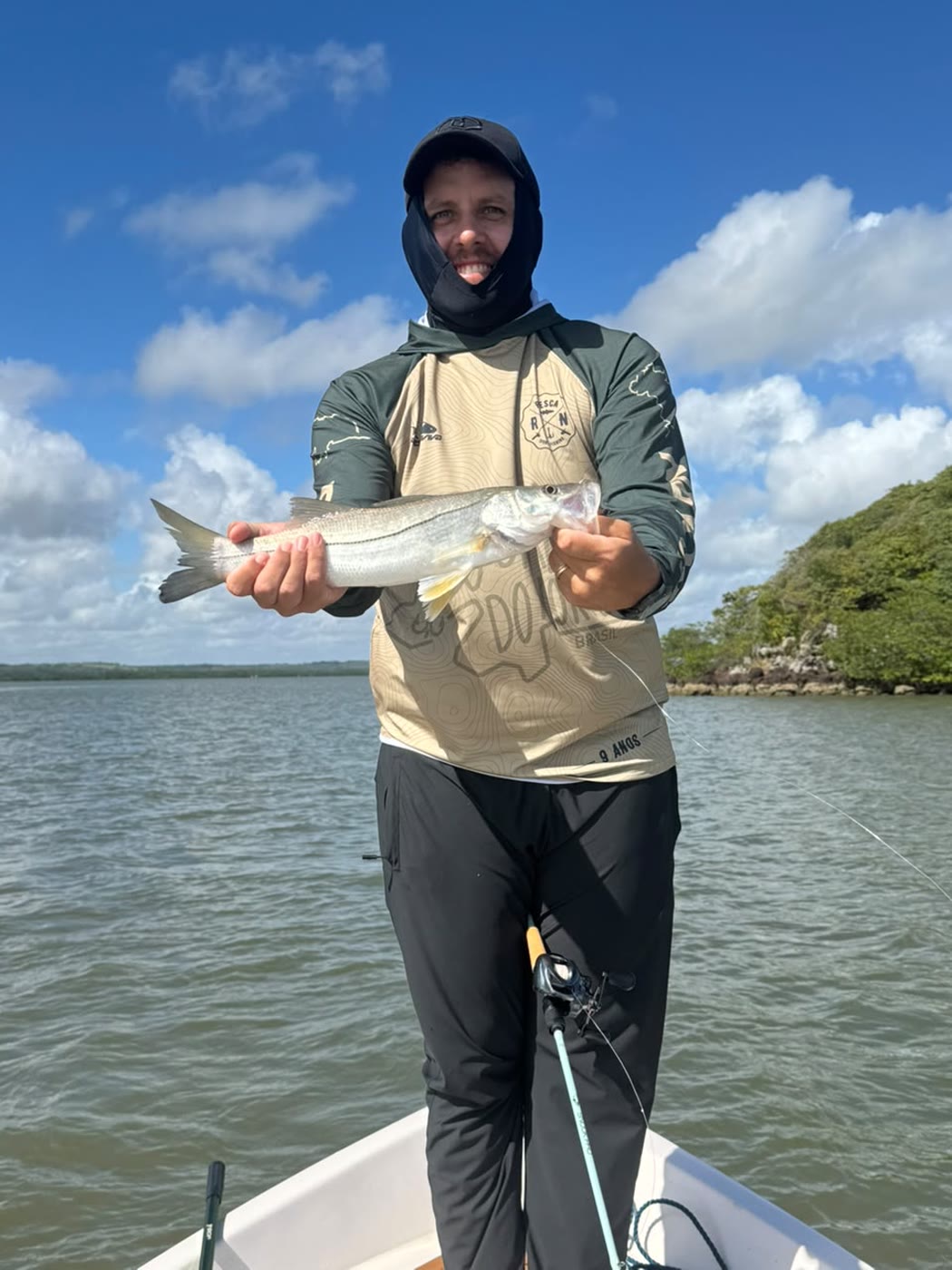 Pescador de touca mostrando o robalo, com mangue ao fundo